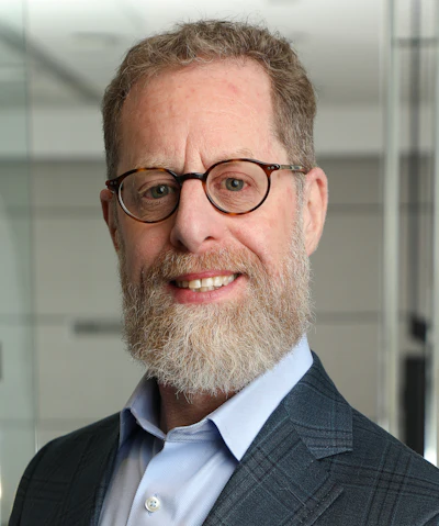 Professional headshot of a bearded man wearing glasses and a blue shirt against a modern office background