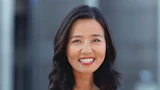 Professional headshot of smiling woman with long dark hair wearing red lipstick against blurred background