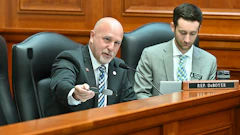 Two legislators seated at a wooden desk during a legislative session, with one speaking into a microphone.