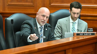 Two legislators seated at a wooden desk during a legislative session, with one speaking into a microphone.