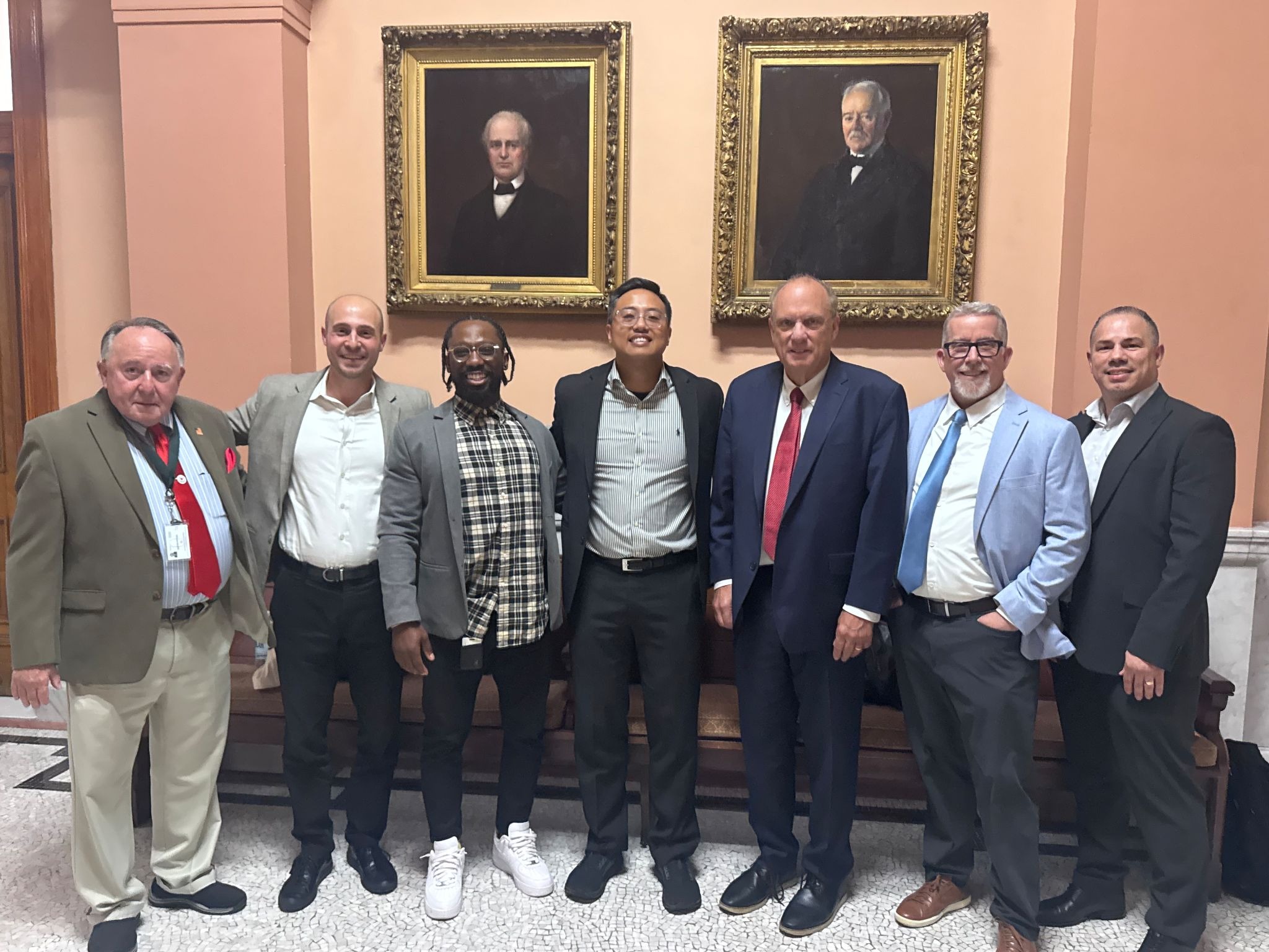 Seven men in business attire posing together in front of ornate framed portraits in a formal institutional setting.