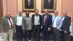 Seven men in business attire posing together in front of ornate framed portraits in a formal institutional setting.