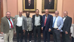 Seven men in business attire posing together in front of ornate framed portraits in a formal institutional setting.