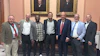 Seven men in business attire posing together in front of ornate framed portraits in a formal institutional setting.