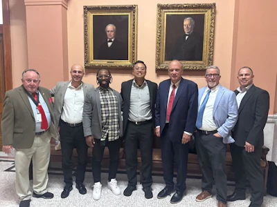 Seven men in business attire posing together in front of ornate framed portraits in a formal institutional setting.