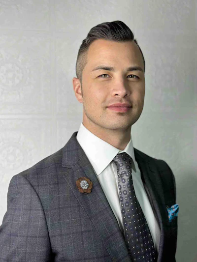 Professional headshot of a man in gray blazer, white dress shirt, and patterned tie against neutral background
