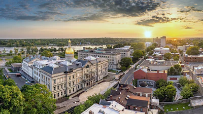Aerial sunset view of downtown with golden-domed state capitol building surrounded by historic architecture and tree-lined streets along a river.