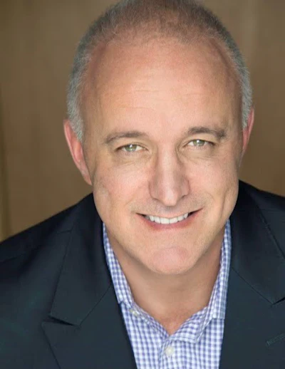 Professional headshot of smiling middle-aged man in dark blazer and blue checkered shirt