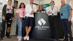 Six team members pose together with a Still River Wellness branded banner at an indoor event