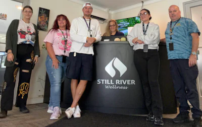 Six team members pose together with a Still River Wellness branded banner at an indoor event